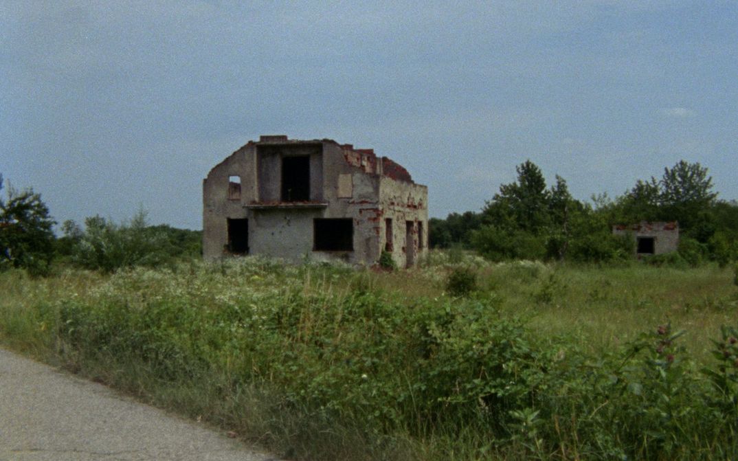 Film still from THOSE SHOCKING SHAKING DAYS. The ruin of a house stands in the middle of an overgrown meadow.