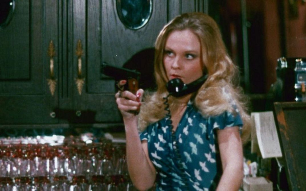 Filmstill aus THE WORKING GIRLS: Eine Frau steht hinter einer Bar und hält eine Pistole in der Hand. Ein Telefonhörer ist zwischen Kopf und Schulter eingeklemmt.