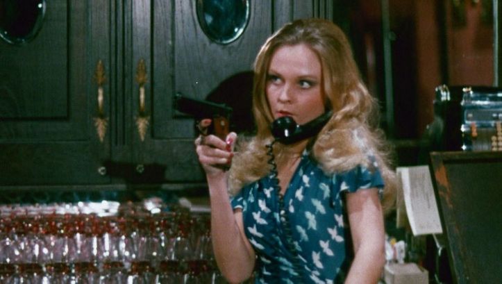 Film still from THE WORKING GIRLS: A woman is standing behind a bar, holding a gun in her hand. A telephone receiver is wedged between her head and shoulder.