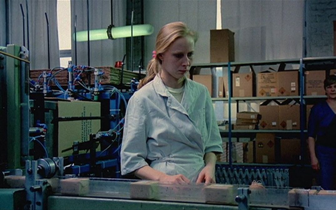 Film still from The Match Factory Girl: A young woman with long blonde hair stands at a factory assembly line, looking distracted.