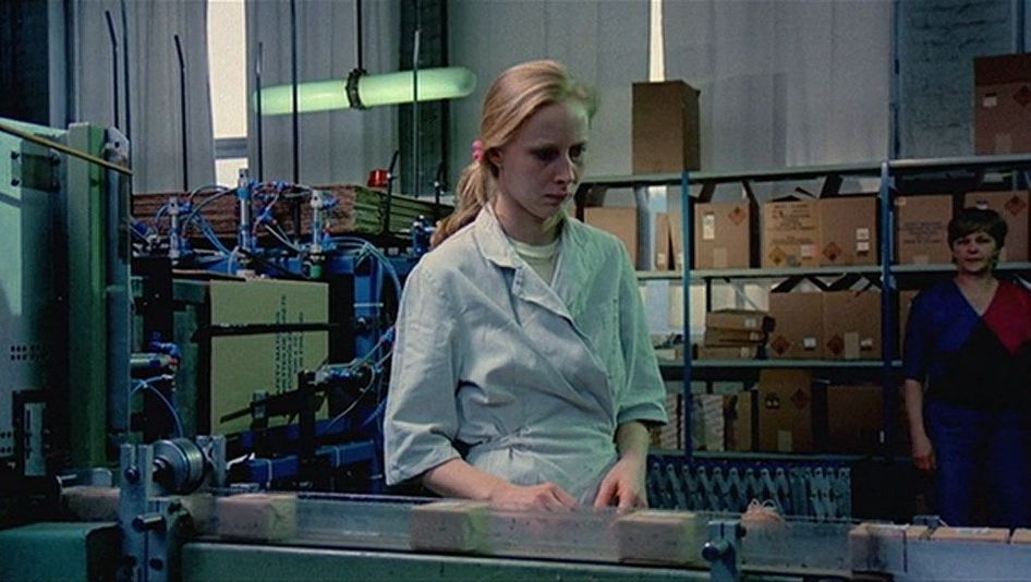 Filmstill aus DAS MÄDCHEN AUS DER STREICHHOLZFABRIK: Eine junge Frau mit langen blonden Haaren steht mit abwesendem Gesichtsausdruck am Fließband einer Fabrik.