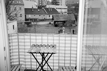Still from the film "Jet Lag" by Zheng Lu Xinyuan. A black-and-white image looking out open glass doors at an ashtray on a wooden table on a balcony, with a backyard and other homes in the background. 