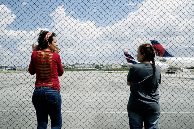 Still from the film "O Estranho" by Flora Dias and Juruna Mallon. Two women stand next to each other in front of a fence and look at each other. Two airplanes standing on an airport runway can be seen in the background.