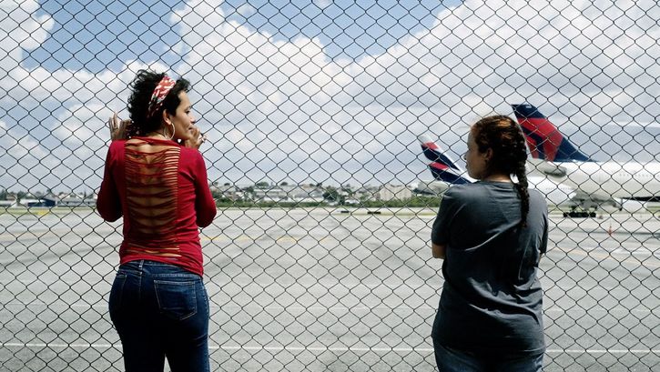 Still from the film "O Estranho" by Flora Dias and Juruna Mallon. Two women stand next to each other in front of a fence and look at each other. Two airplanes standing on an airport runway can be seen in the background.
