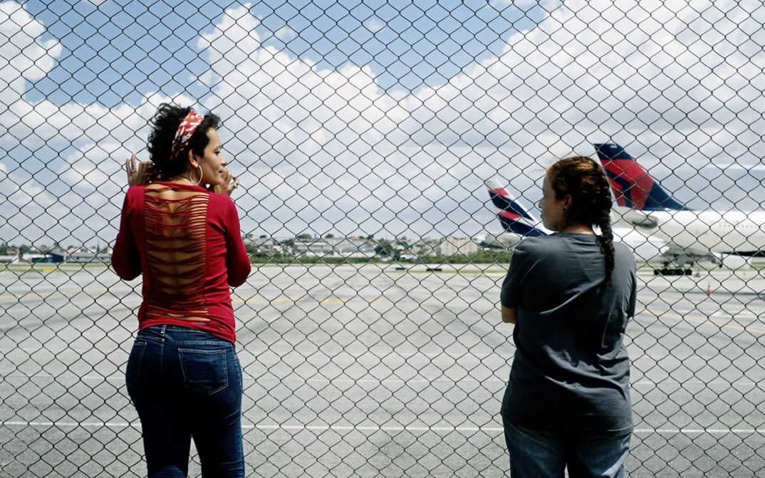 Filmstill aus dem Film "O Estranho" von Flora Dias und Juruna Mallon. Zwei Frauen stehen nebeneinander vor einem Zaun und schauen sich an, im Hintergrund sind zwei Flugzeuge auf einer Landebahn zu sehen.
