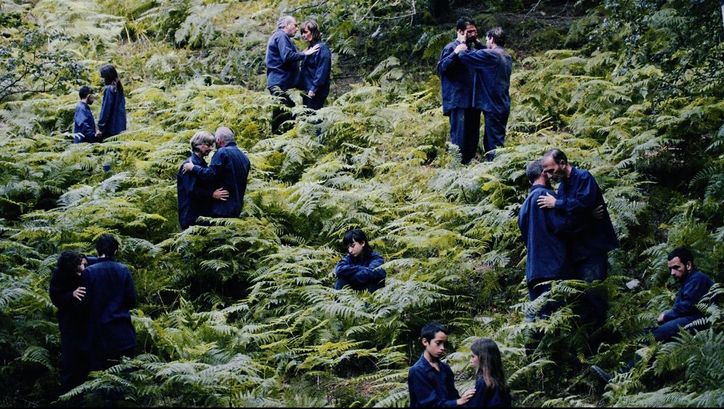 Film still from "The Human Hibernation" by Anna Cornudella Castro.It shows people of different ages spread out in pairs in a field of ferns. One person is sitting alone. They are all wearing dark blue clothing.