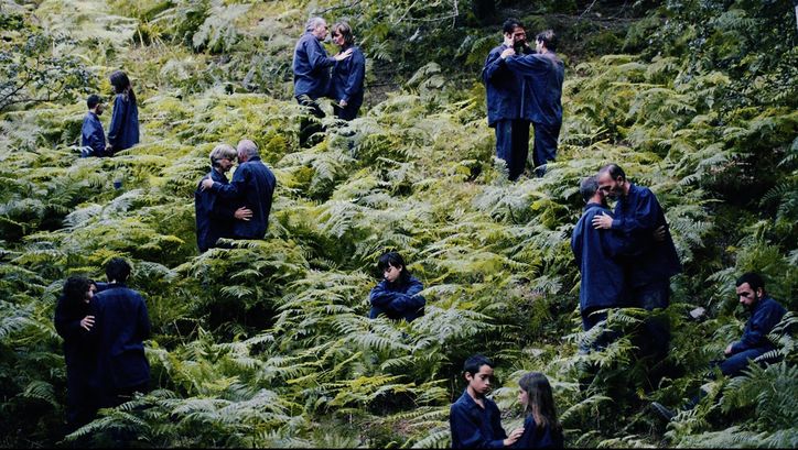 Filmstill aus "The Human Hibernation" von Anna Cornudella Castro. Zu sehen sind Personen unterschiedlichen Alters, die paarweise in einem Farnfeld verteilt sind. Eine Person sitzt allein. Sie tragen alle dunkelblaue Kleidung. 