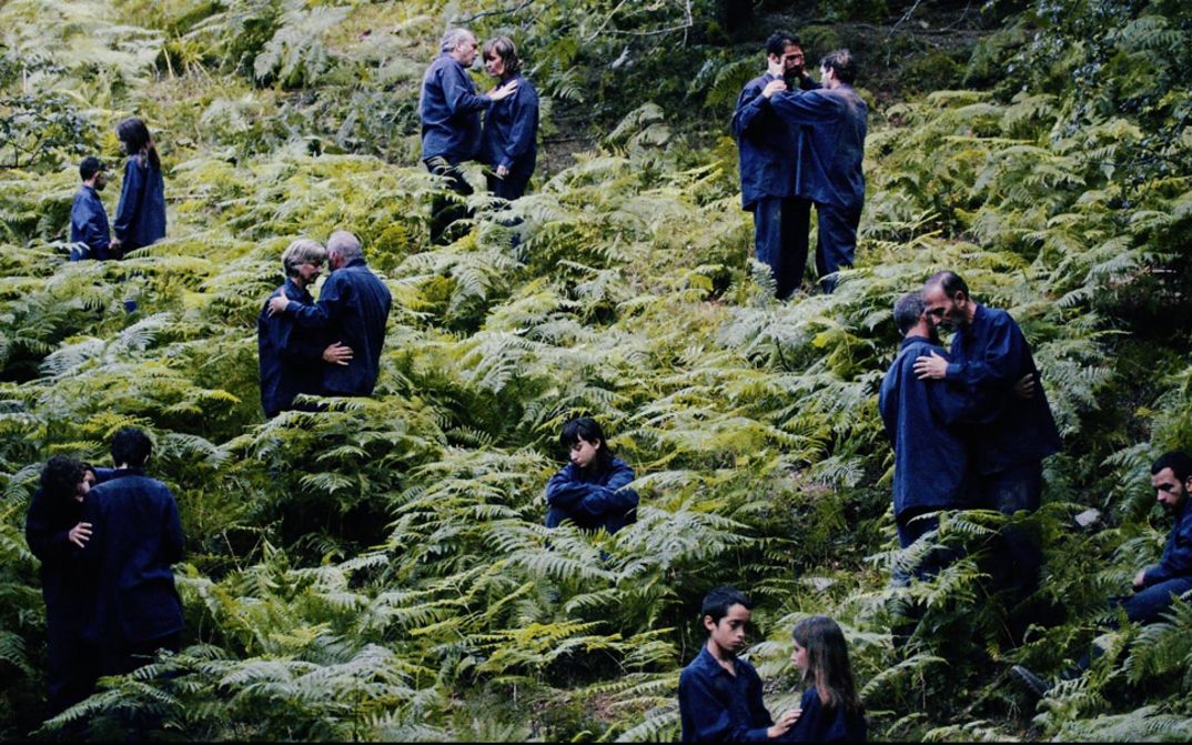 Film still from "The Human Hibernation" by Anna Cornudella Castro.It shows people of different ages spread out in pairs in a field of ferns. One person is sitting alone. They are all wearing dark blue clothing.