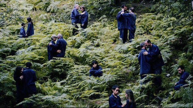 Film still from "The Human Hibernation" by Anna Cornudella Castro.It shows people of different ages spread out in pairs in a field of ferns. One person is sitting alone. They are all wearing dark blue clothing.