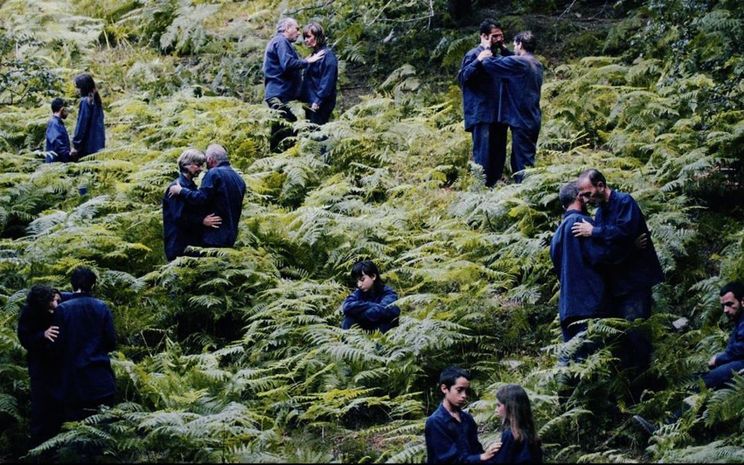 Filmstill aus "The Human Hibernation" von Anna Cornudella Castro. Zu sehen sind Personen unterschiedlichen Alters, die paarweise in einem Farnfeld verteilt sind. Eine Person sitzt allein. Sie tragen alle dunkelblaue Kleidung.