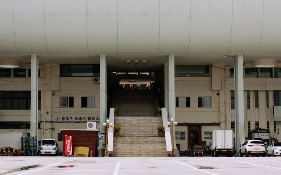 Film still from THE SENSE OF VIOLENCE by Mooyung Kim. The picture shows the ground floor of a large building. Cars are parked to the left and right of a staircase.