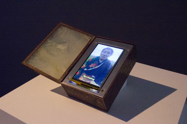 Installation view of Tenzin Phuntsog’s „Achala“. An opened box with a screen inside. On the screen a person in a robe with a flower pattern.
