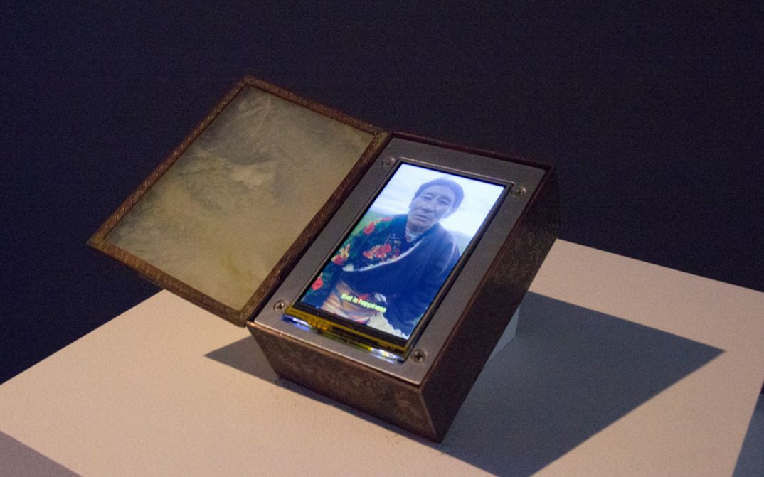 Installation view of Tenzin Phuntsog’s „Achala“. An opened box with a screen inside. On the screen a person in a robe with a flower pattern.