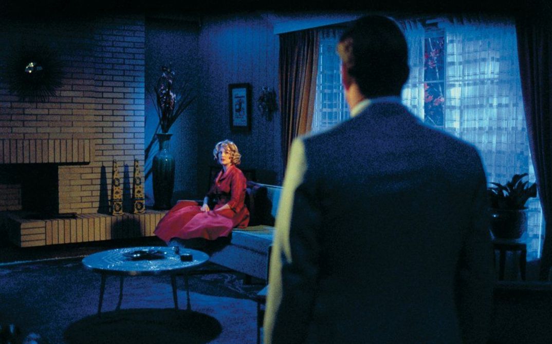 Filmstill from FAR FROM HEAVEN: A woman in a red dress is sitting in a living room. It is dark. A man can be seen from behind, looking in her direction.