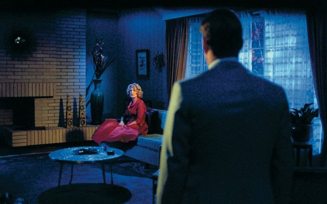 Filmstill aus FAR FROM HEAVEN: Eine Frau in einem roten Kleid sitzt in einem Wohnzimmer. Es ist dunkel. Ein Mann ist von hinten zu sehen, der in ihre Richtung blickt.
