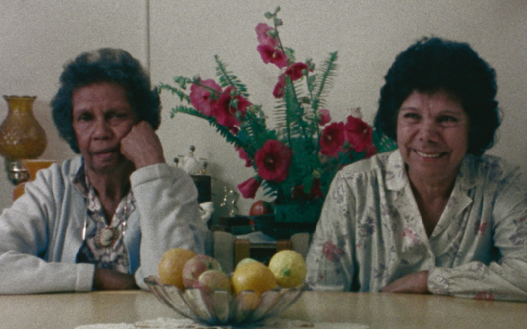 Filmstill aus LOUSY LITTLE SIXPENCE: Zwei Frauen sitzen an einem Tisch und blicken in die Kamera. Hinter ihnen ist ein Blumengedeck.