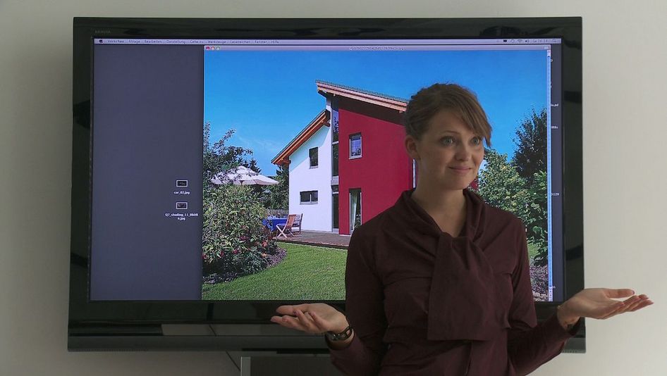 Filmstill aus EINE FELXIBLE FRAU: Eine Frau steht im Modus des Präsentierens vor einem großem Monitor, auf dem ein Einfamilienhaus zu sehen ist. 