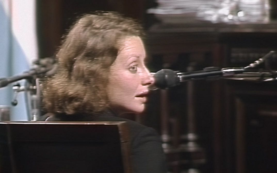 Filmstill aus dem Film "El juicio" von Ulises de la Orden. Eine Frau sitzt in einem Zeugenstand und schaut zu ihrer rechten, neben ihr steht ein Mikrofon.