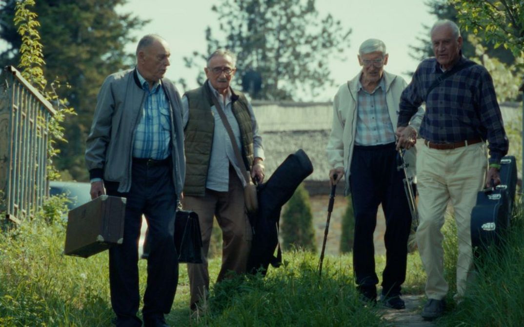 Filmstill aus RESTITUCIJA, ILI, SAN I JAVA STARE Gare von Želimir Žilnik. Das Bild zeigt vier ältere Herren mit Koffern und Instrumententaschen im Freien.