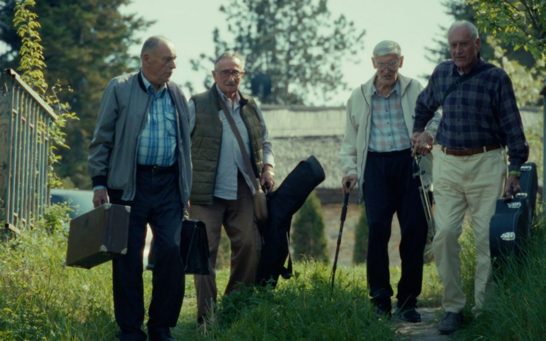 Filmstill aus RESTITUCIJA, ILI, SAN I JAVA STARE Gare von Želimir Žilnik. Das Bild zeigt vier ältere Herren mit Koffern und Instrumententaschen im Freien.