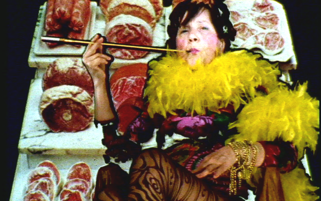 Filmstill aus MARIA LASSNIG KANTATE: Zu sehen ist eine auffällig gekleidete Person mit einer Federboa und einer Zigarettenspitze. Der Hintergrund besteht aus Thekenfleisch.