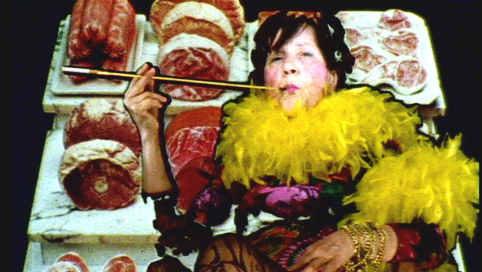 Film still from MARIA LASSNIG KANTATE: It shows a strikingly dressed person with a feather boa and a cigarette holder. The background consists of counter meat.