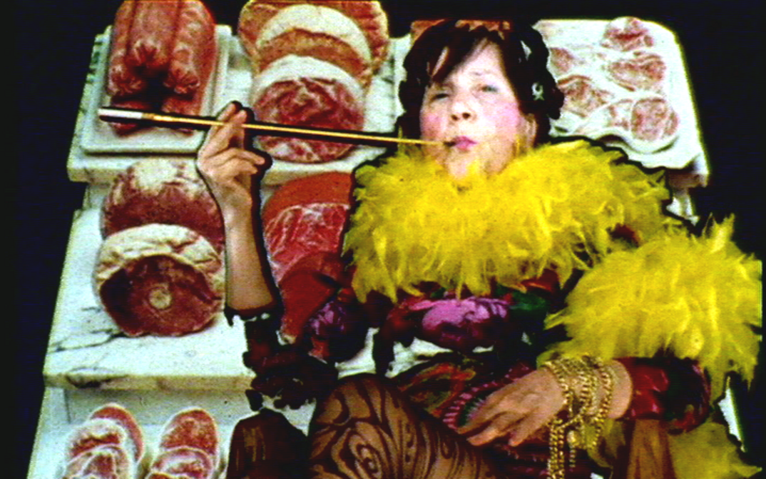 Film still from MARIA LASSNIG KANTATE: It shows a strikingly dressed person with a feather boa and a cigarette holder. The background consists of counter meat.