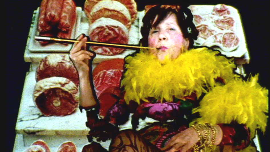 Filmstill aus MARIA LASSNIG KANTATE: Zu sehen ist eine auffällig gekleidete Person mit einer Federboa und einer Zigarettenspitze. Der Hintergrund besteht aus Thekenfleisch.