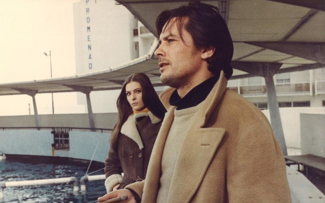 Film still from LA PRIMA NOTTE DI QUIETE: Alain Delon is standing by a railing by the sea, wearing a coat. A young woman approaches him.