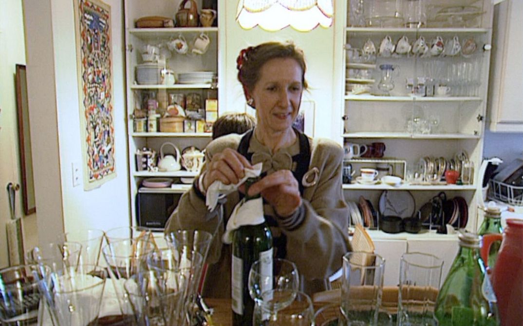 Filmstill from MANHATTAN STORIES: A woman in an apron wipes a wine bottle. Around her are many glasses and kitchen utensils.