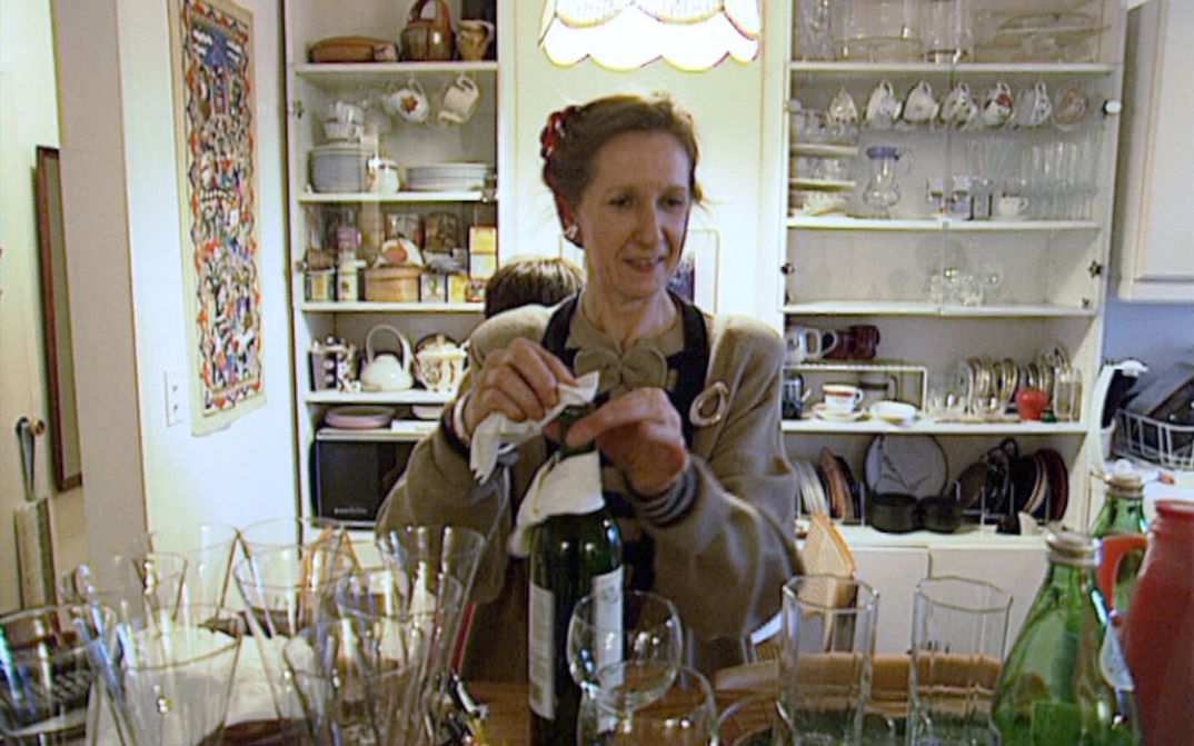 Filmstill from MANHATTAN STORIES: A woman in an apron wipes a wine bottle. Around her are many glasses and kitchen utensils.