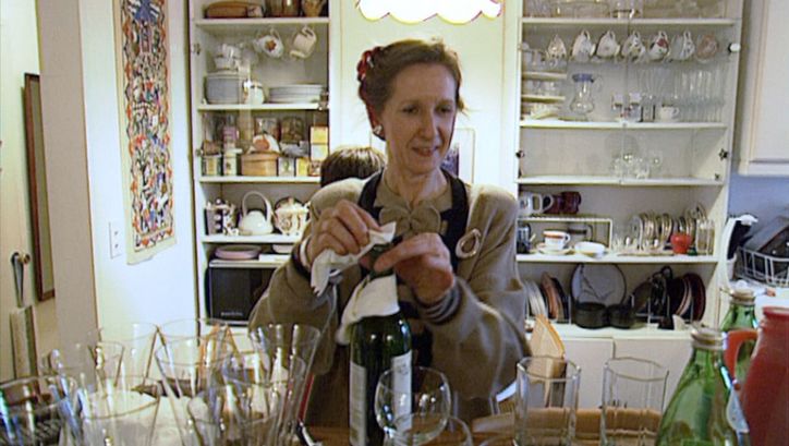 Filmstill from MANHATTAN STORIES: A woman in an apron wipes a wine bottle. Around her are many glasses and kitchen utensils.
