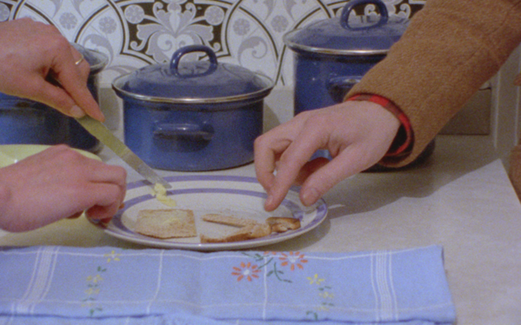 Film still from RIDDLES OF THE SPHINX: On a table are three blue pots and a floral kitchen towel. In the middle is a plate with a piece of bread; two hands are reaching for it.