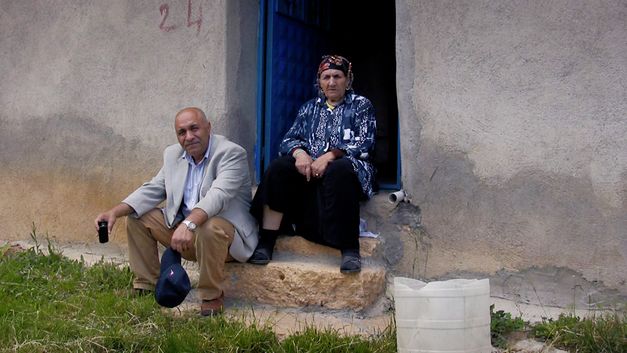 Still from the film "Dilim dönmüyor – Meine Zunge dreht sich nicht" by Serpil Turhan. An older man and a woman in village clothing sit on the stairs in front of their house. 