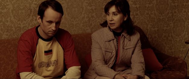 Film still from JANINE ZIEHT AUFS LAND by Jan Eilhardt. The picture shows a woman and a man on a sofa. The woman is talking to the man, he is looking at the floor.
