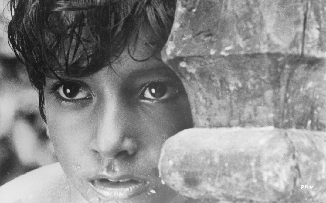 Filmstill aus PATHER PANCHALI: Ein Junge wirkt, als würde er etwas beobachten und sich hinter einer Säule oder Holz verstecken.