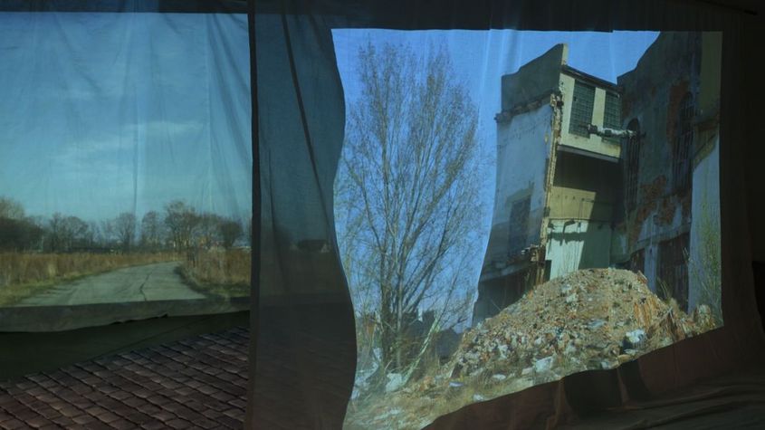 Filmstill aus dem Film "The Song of the Shirt" von Kerstin Schroedinger. Durch transparenten Stoff erhält man einen blick auf ein zerfallenes Haus.