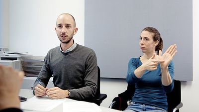 Still from the film "For the Many – The Vienna Chamber of Labour" by Constantin Wulff: A man is sitting in an office, speaking to someone off camera. To the right, a woman is translating to sign language.