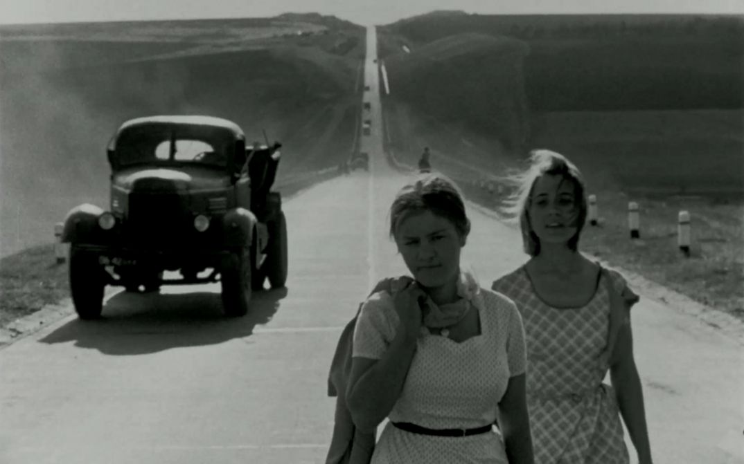 Filmstill auf BRIEF ENCOUNTERS: Zwei junge, sommerlich gekleidete Frauen gehen eine Straße entlang. Hinter ihnen ist ein Lastwagen zu sehen.