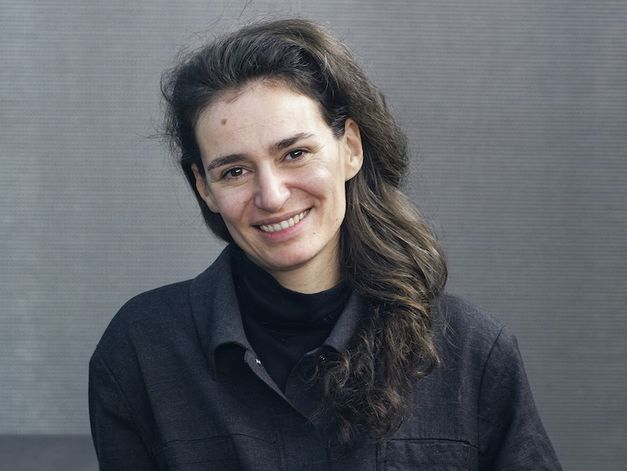 Marta Mateus poses in front of a grey wall. Her hands are clasped and her right arm rest on a counter.