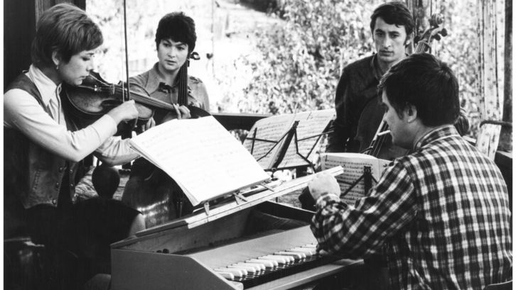 Film still from PASTORALE: A group of musicians stand around a piano.