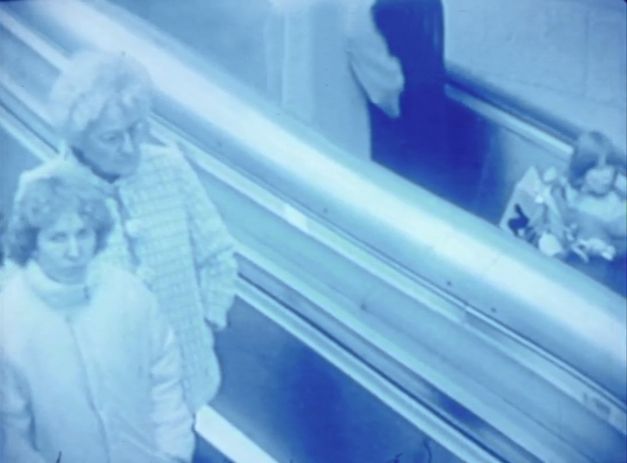 Filmstill aus „Merry Christmas Deutschland oder Vorlesung zur Geschichtstheorie II“ von Raoul Peck. Ein blau eingefärbtes Bild von Menschen, die auf einer Rolltreppe fahren. 