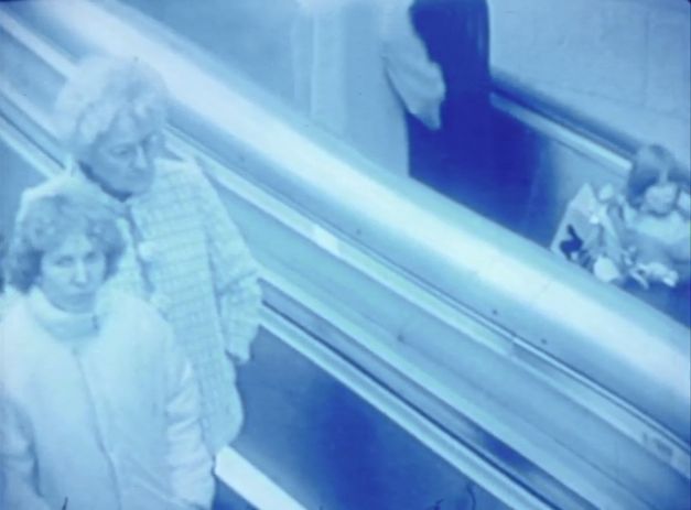 Still from the film "Merry Christmas Deutschland oder Vorlesung zur Geschichtstheorie II" by Raoul Peck. A blue tinted image of people riding an escalator. 