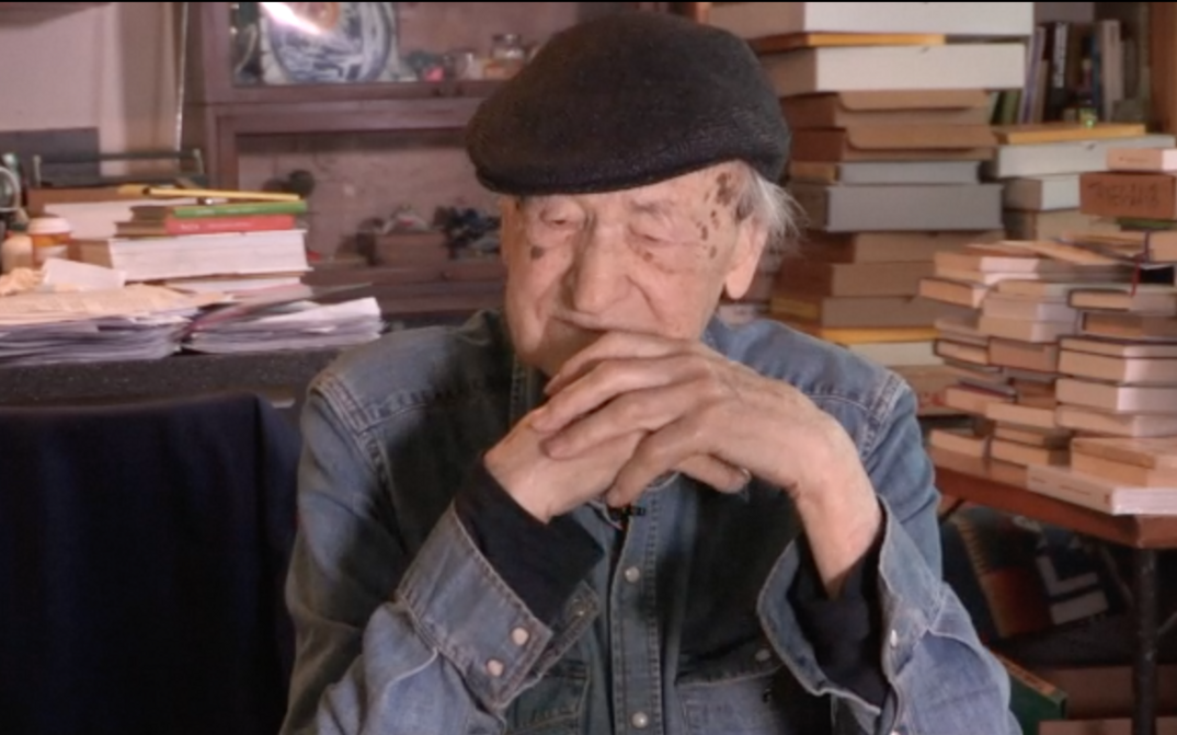 Film still from ORAL HISTORY INTERVIEW: JONAS MEKAS: Jonas Mekas sitting, tables full of books behind him.