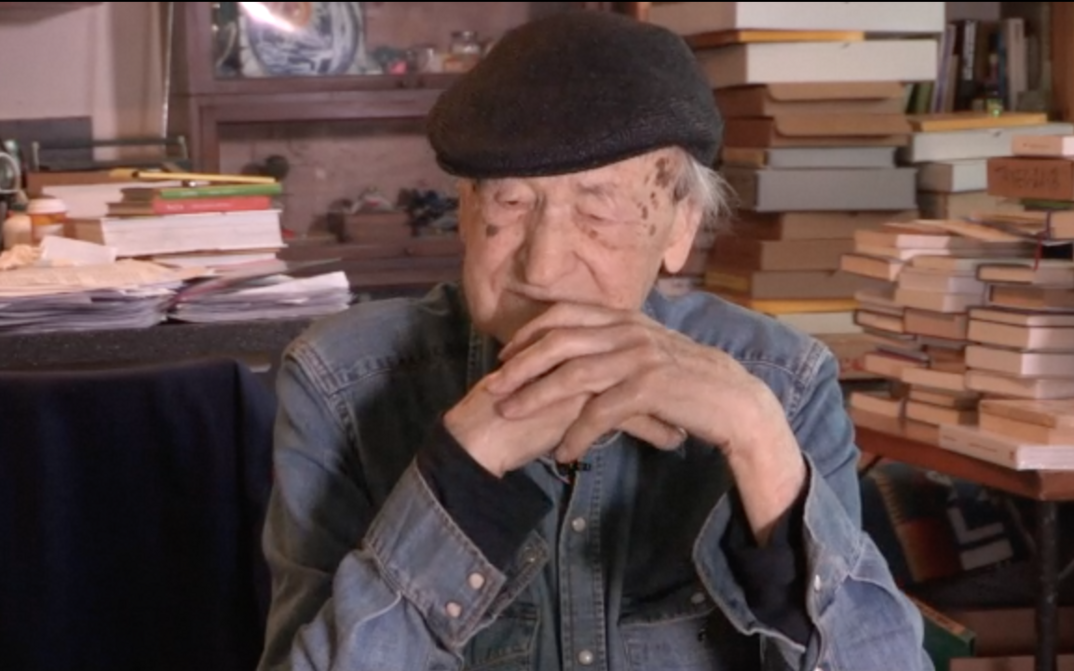 Filmstill aus ORAL HISTORY INTERVIEW: JONAS MEKAS: Jonas Mekas sitztend, hinter ihm Tische voller Bücher.