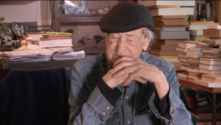 Film still from ORAL HISTORY INTERVIEW: JONAS MEKAS: Jonas Mekas sitting, tables full of books behind him.