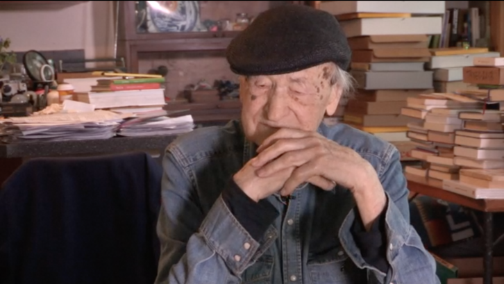 Film still from ORAL HISTORY INTERVIEW: JONAS MEKAS: Jonas Mekas sitting, tables full of books behind him.