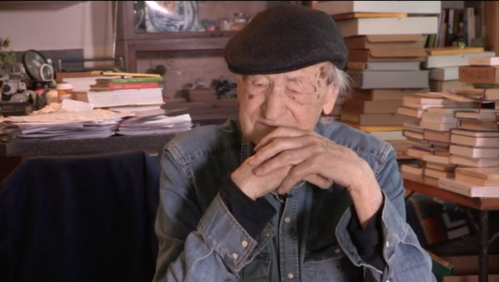 Filmstill aus ORAL HISTORY INTERVIEW: JONAS MEKAS: Jonas Mekas sitztend, hinter ihm Tische voller Bücher.