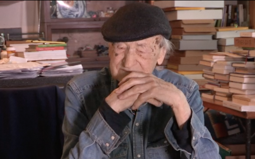Film still from ORAL HISTORY INTERVIEW: JONAS MEKAS: Jonas Mekas sitting, tables full of books behind him.