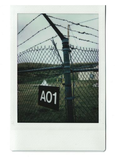 Polaroid from the outside of a chain-linked fence with a sign reading A01. Behind we see green fields and a house.